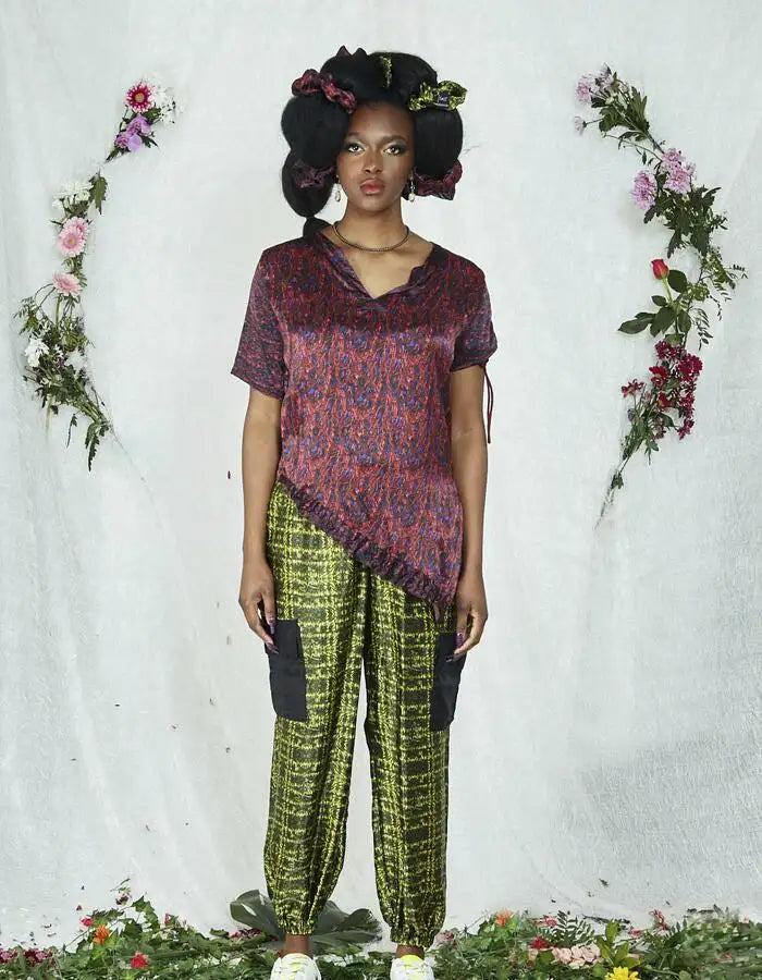 Model in asymmetrical red and purple print top paired with lime green check cargo trousers, styled with voluminous hair adorned with floral scrunchies, standing in front of a soft white backdrop decorated with flowers