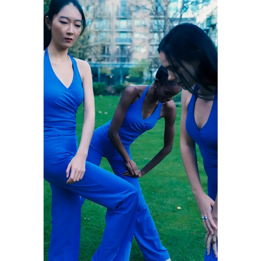 Three women in blue outfits standing on a grassy area with trees and buildings in the background.