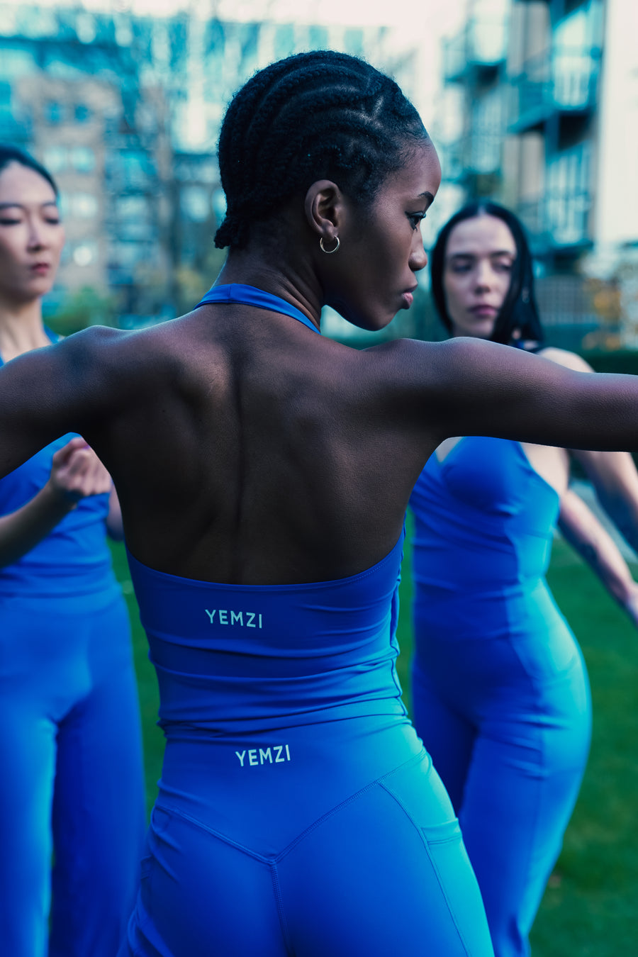 Three women in blue YEMZI outfits standing outdoors with a cityscape background.