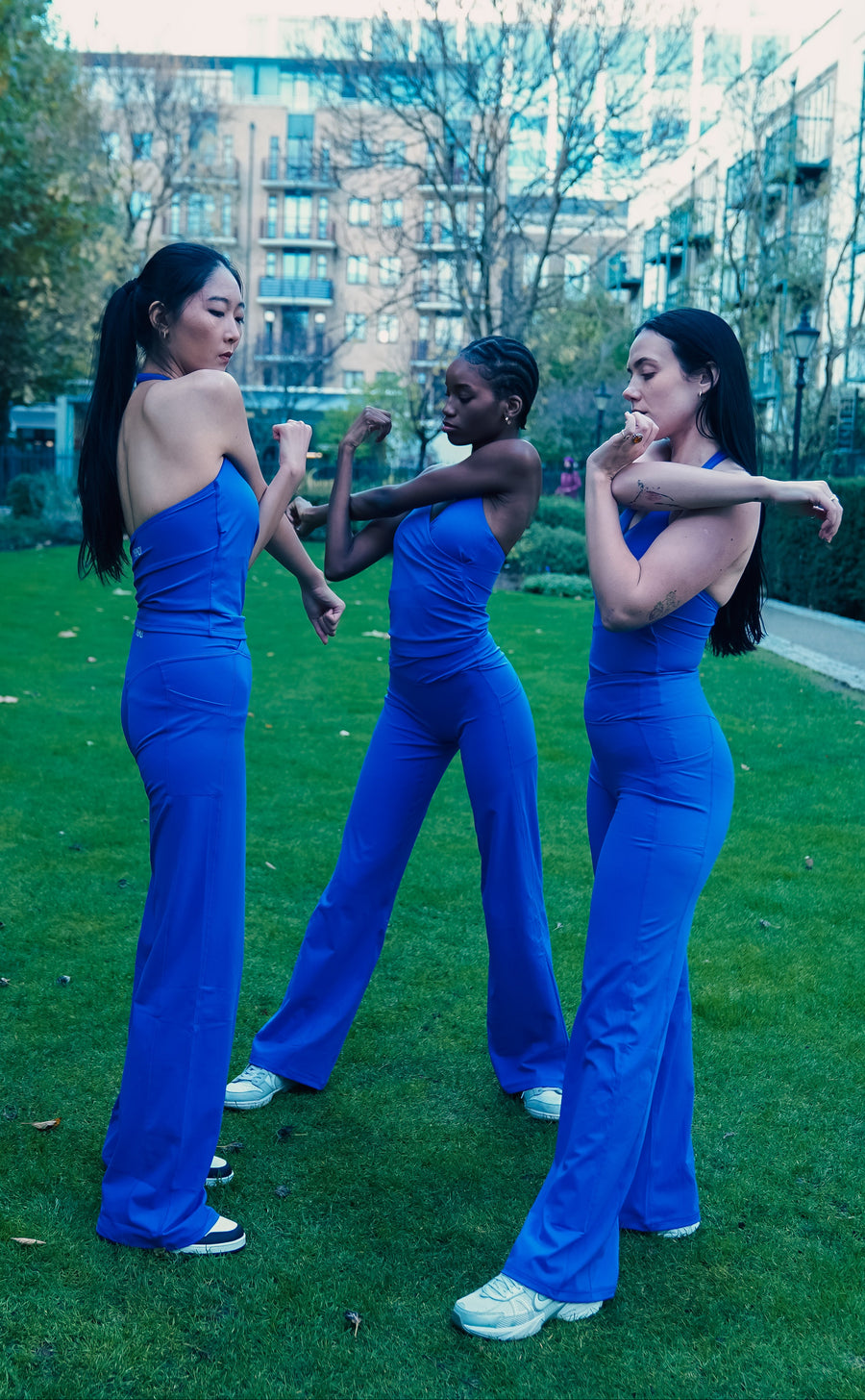 Three women in matching blue outfits standing on a grassy area with buildings in the background.