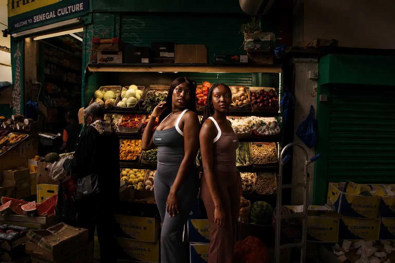 Two women stand side by side in front of a vibrant green fruit and vegetable stall, one wearing a gray tank top with white trim paired with matching leggings, the other in a brown bodysuit.